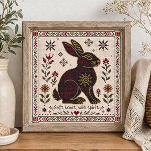May include: A framed cross-stitch featuring a black rabbit with burgundy and gold accents. The design includes floral and snowflake motifs, with the text "Soft heart, wild spirit." The artwork is set against a cream background and framed in a light brown wooden frame.