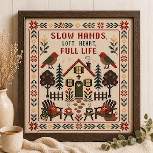 Op de afbeelding: Kruissteekkunst in een donkerhouten lijst. Het ontwerp toont een huis, bomen, vogels en de tekst "SLOW HANDS, SOFT HEART, FULL LIFE." Het kleurenpalet omvat rood, groen en bruin, wat een gezellige, rustieke sfeer creëert.