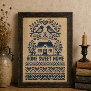 Może przedstawiać: Oprawiony haft krzyżykowy na beżowej tkaninie z ciemnoniebieskim wzorem przedstawiającym dwa ptaki, serce, dom oraz napis HOME SWEET HOME, a poniżej wzór geometryczny. Całość umieszczona jest w ciemnej, postarzanej drewnianej ramie.