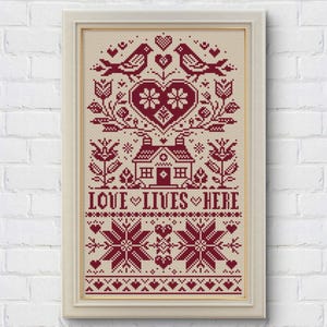 May include: Framed cross-stitch artwork with a cream background and a burgundy design. The design features birds, hearts, flowers, a house, and the text "LOVE LIVES HERE". The artwork is in a white frame and is displayed against a white brick wall.