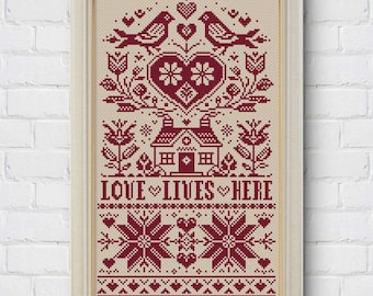 Puede incluir: Obra de arte en punto de cruz enmarcada con un fondo crema y un diseño burdeos. El diseño presenta pájaros, corazones, flores, una casa y el texto "LOVE LIVES HERE". La obra de arte está en un marco blanco y se exhibe contra una pared de ladrillos blancos.