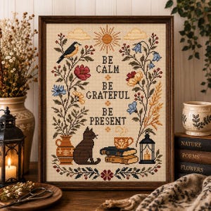 Peut inclure: Une broderie au point de croix encadrée avec le texte BE CALM BE GRATEFUL BE PRESENT. Le motif comprend un soleil, des nuages, des fleurs, un oiseau, un chat noir, une pile de livres avec une tasse de thé et une lanterne, brodés en couleurs.