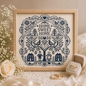 May include: A framed cross-stitch sampler with the words "HOME SWEET HOME" in navy blue. The design includes birds, trees, houses, and a couple. A wedding ring set and a ring box are in the foreground. The frame is light brown.