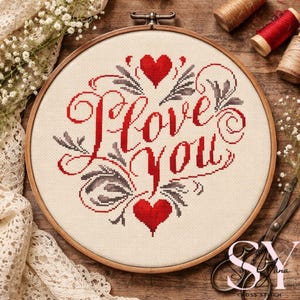以下が含まれることがあります： 赤い文字で「I Love You」と書かれたクロスステッチ刺繍枠。赤いハートと灰色の花のアクセントで囲まれています。刺繍はクリーム色の生地に施され、木製の枠で縁取られています。