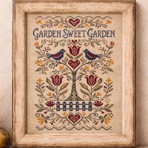 May include: Framed cross-stitch artwork with a rustic wooden frame. The design features a tree with birds, hearts, and floral elements in shades of red, blue, and yellow. The text "GARDEN SWEET GARDEN" is stitched across the top.