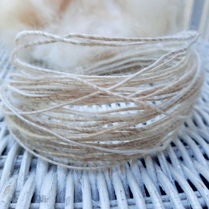 May include: A close-up of a skein of natural fiber yarn in a light beige color. The yarn is loosely wound and has a soft, fuzzy texture.