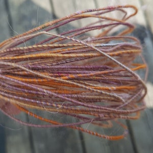 Puede incluir: Un ovillo de hilo con una mezcla de colores naranja, marrón y morado. El hilo es suave y esponjoso, y sería perfecto para tejer o hacer ganchillo.