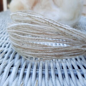 May include: Close-up of a bundle of natural fiber yarn, featuring a mix of white and beige strands. The yarn is loosely wound and has a soft, fluffy texture.