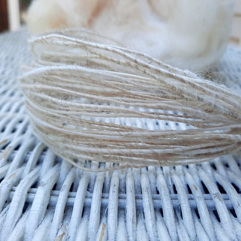 May include: Close-up of a bundle of natural fiber yarn, featuring a mix of white and beige strands. The yarn is loosely wound and has a soft, fluffy texture.