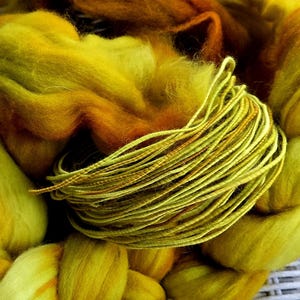 Puede incluir: Un manojo enrollado de cordón fino de color verde lima que descansa sobre una gruesa masa trenzada de lana cardada sin hilar. La lana presenta un degradado de tonos amarillo vibrante, naranja dorado y ámbar intenso, creando una muestra de fibras texturizada y multicolor.