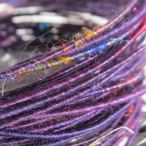 May include: Close-up of a spool of purple yarn with red, blue, and gold accents. The yarn is tightly wound and has a fuzzy texture.