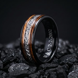 May include: A black ring featuring a central inlay of textured silver-toned metallic material flanked by two bands of brown wood. The interior of the band is engraved with the white text B & C 2-7-2002, showcasing a polished finish.
