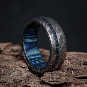 May include: A men's damascus steel ring with a blue and white wood inlay. The ring features a unique pattern on the metal band and a contrasting wood insert. The image showcases the intricate details of the craftsmanship and the rich colors of the materials. This handcrafted ring is a stylish accessory for men.