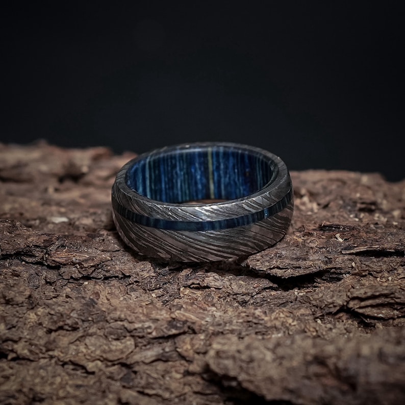 May include: A silver ring with a blue wood inlay. The ring has a hammered finish and a wide band.