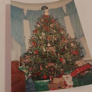 May include: A decorated Christmas tree with baubles, fairy lights, and presents underneath. The tree is in a living room with blue curtains and a brown armchair.