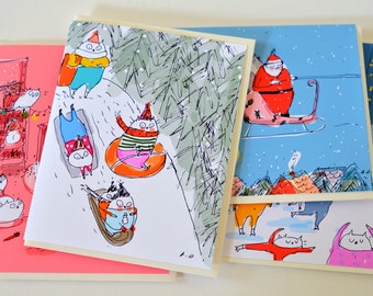May include: Five illustrated Christmas cards featuring cats in winter scenes. One card shows a cat wearing a red Santa hat and riding a sled pulled by a reindeer. Another card shows a cat wearing a red and white striped sweater and riding a snow tube. Another card shows a cat wearing a blue sweater and riding a sled. Another card shows a cat wearing a pink sweater and riding a sled. The last card shows a cat wearing a pink sweater and riding a snowboard.