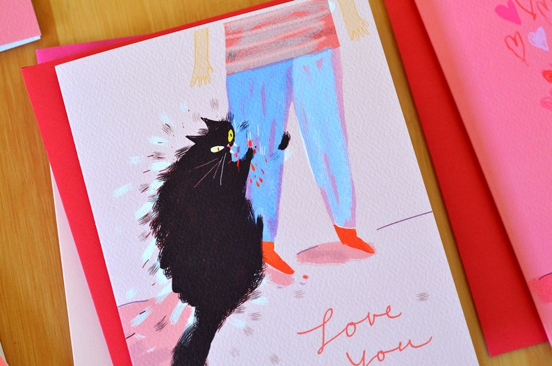 May include: A pink greeting card with a black cat scratching a person's leg. The person is wearing blue pants and red shoes. The card says "love you" in pink cursive writing.