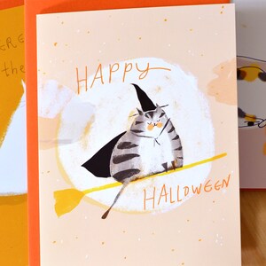 May include: A greeting card with a cute illustration of a cat wearing a witch's hat and riding a broomstick. The card has a peach background with orange and white speckles. The text "Happy Halloween" is written in black on the card.