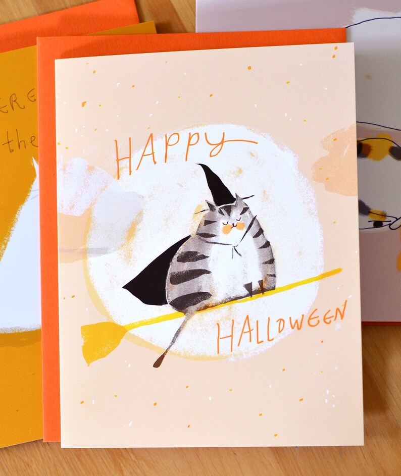 May include: A greeting card with a cute illustration of a cat wearing a witch's hat and riding a broomstick. The card has a peach background with orange and white speckles. The text "Happy Halloween" is written in black on the card.