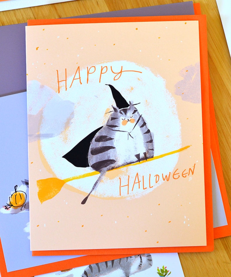 May include: A greeting card with a peach background and orange border. The card features a cartoon illustration of a gray and white cat wearing a witch's hat and cape, riding a yellow broomstick. The text "HAPPY HALLOWEEN" is written in black on the card.