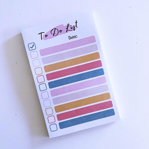 May include: A white notepad with the words "To Do List" in black script and a pink watercolour wash. The notepad has a checklist with colourful horizontal bars in shades of pink, blue, and gold. The word "Date:" is printed below the title.
