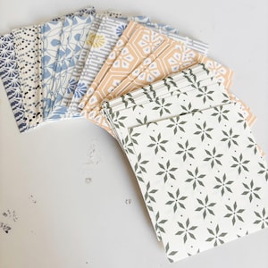 May include: A selection of patterned paper sheets with diverse designs. The patterns feature floral, geometric, and abstract motifs in blue, yellow, tan, and green. The sheets are arranged in a fan shape on a white surface.