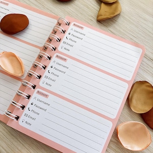 May include: A pink and white spiral-bound notebook with sections for account details. Each section includes fields for username, password, phone, email, and notes. Decorative items are scattered around the notebook.