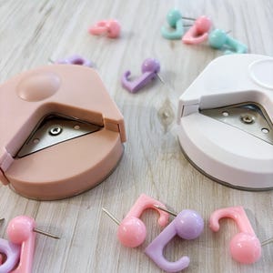 May include: Two round corner punch tools, one pink and one white, with a silver blade. The tools are on a light wood surface with several colourful push pins with hooks.