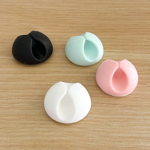 May include: Four cable organisers in different colours: black, light blue, pink, and white. Each organiser has a round shape with a hole in the centre for holding cables.