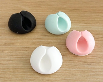 May include: Four cable organizers in different colors: black, light blue, pink, and white. Each organizer has a round shape with a hole in the center for holding cables.