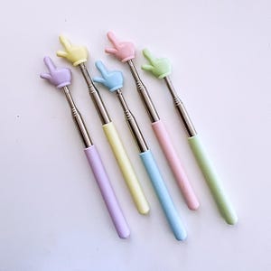 K&ouml;nnte beinhalten: F&uuml;nf einziehbare Selfie-Sticks mit pastellfarbenen Griffen in Form von H&auml;nden, die ein Peace-Zeichen machen. Die Griffe sind lavendelfarben, gelb, hellblau, rosa und hellgr&uuml;n.