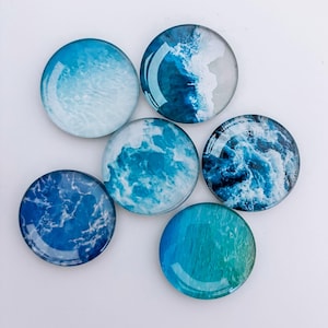 May include: Six circular glass magnets featuring ocean wave patterns in various shades of blue and white. The designs range from gentle to rough waves. The glass has a glossy finish.