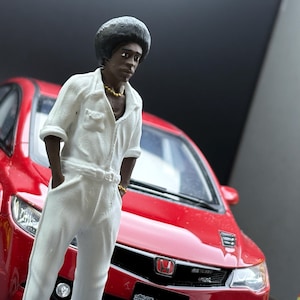 May include: A miniature figure of a person in a white jumpsuit, gold necklace, and bracelet stands next to a red sports car. The figure has a dark complexion and a black afro. The car has black rims and a black front grill.