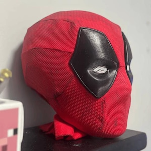 May include: A red Deadpool mask with black eye coverings and white mesh eye inserts. The mask is made of a textured fabric and sits on a black surface. The mask is a cosplay item.