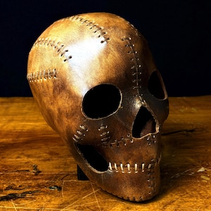 Pode incluir: Uma máscara de caveira de couro castanho com costuras visíveis. A máscara tem aberturas para os olhos e o nariz. As costuras são de uma cor mais clara, criando contraste. Exibida sobre uma superfície de madeira.