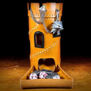 May include: A tan leather dice tower in a castle design. The tower has a silver chain with a wolf pendant and a tray at the base holding dice. The tower has a stitched design and small windows. The background is dark.