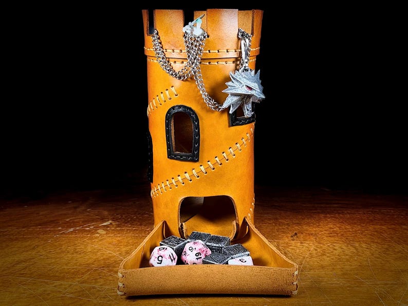 May include: A tan leather dice tower in a castle design. The tower has a silver chain with a wolf pendant and a tray at the base holding dice. The tower has a stitched design and small windows. The background is dark.