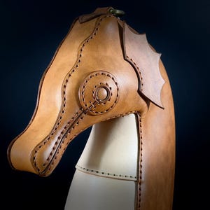 May include: A handcrafted leather seahorse sculpture in shades of tan and cream. The seahorse head is detailed with stitched accents and a circular design. The sculpture has a dark background.