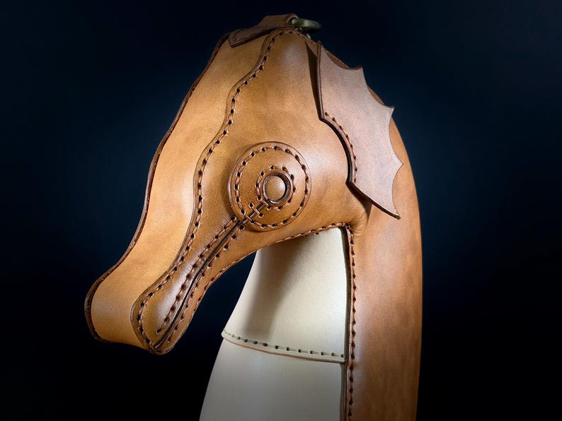 May include: A handcrafted leather seahorse sculpture in shades of tan and cream. The seahorse head is detailed with stitched accents and a circular design. The sculpture has a dark background.