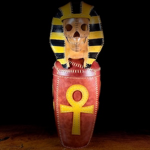 Könnte beinhalten: Handgefertigte Lederskulptur eines ägyptischen Pharaonensarkophags. Die Skulptur zeigt ein Totenkopfgesicht, eine gelb-schwarz gestreifte Kopfbedeckung und einen roten Körper mit einem gelben Ankh-Symbol. Das Stück ist mit weißem Faden zusammengenäht.