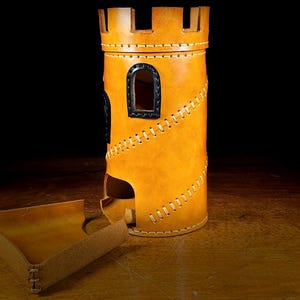 May include: A tan leather dice tower, designed to resemble a castle. It features black window accents and decorative stitching. A matching tray is positioned beside the tower. The tower is cylindrical with a slot for dice.