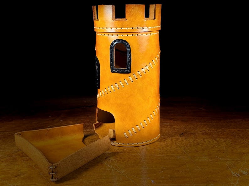 May include: A tan leather dice tower, designed to resemble a castle. It features black window accents and decorative stitching. A matching tray is positioned beside the tower. The tower is cylindrical with a slot for dice.