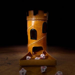 May include: A handcrafted, tan leather dice tower in the shape of a castle. The tower has two arched windows and a stitched design. A tray is attached to the base, with several dice visible. The background is dark, and the tower sits on a wooden surface.