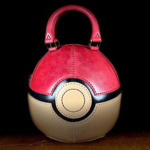 Op de afbeelding: Een rode, zwarte en bruin/beige handtas met een ronde vorm, die lijkt op een Poké Ball. De tas heeft een rode handgreep en goudkleurige hardware. De tas is gemaakt van gestikt leer en heeft een cirkelvormig ontwerp aan de voorkant.