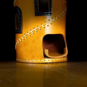 May include: A tan leather dice tower with a cylindrical shape. It has a rectangular opening and two small window-like cutouts with black borders. The seams are stitched with yellow thread, creating a decorative pattern. The tower is designed for tabletop gaming.