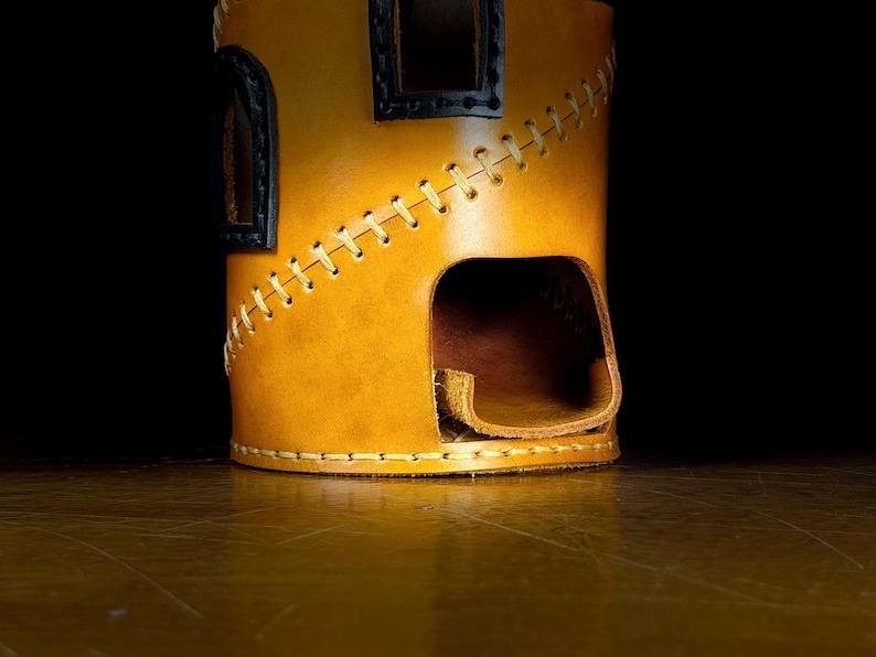 May include: A tan leather dice tower with a cylindrical shape. It has a rectangular opening and two small window-like cutouts with black borders. The seams are stitched with yellow thread, creating a decorative pattern. The tower is designed for tabletop gaming.