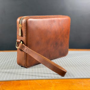 May include: A rectangular brown leather clutch bag featuring a smooth finish, visible stitched edges, and a gold-toned metal zipper closure. The bag includes a matching leather wrist strap attached to a gold-toned metal D-ring on the side.