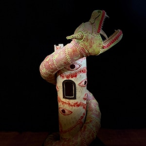 Può includere: Un'originale scultura di un serpente avvolto attorno a una struttura cilindrica. La testa del serpente è aperta, rivelando i denti. La scultura è principalmente color crema con dettagli rossi e verdi e presenta molteplici disegni di occhi.