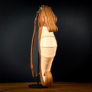 May include: A handcrafted leather seahorse-shaped bag in tan and cream colors. The bag features detailed stitching and a long adjustable strap. The seahorse design includes a head, body, and tail, with a unique, artistic aesthetic.