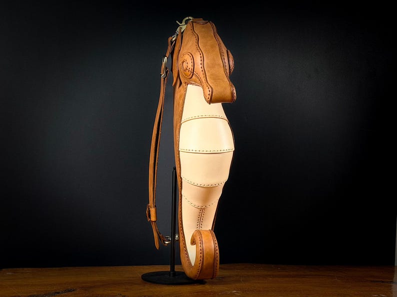 May include: A handcrafted leather seahorse-shaped bag in tan and cream colors. The bag features detailed stitching and a long adjustable strap. The seahorse design includes a head, body, and tail, with a unique, artistic aesthetic.